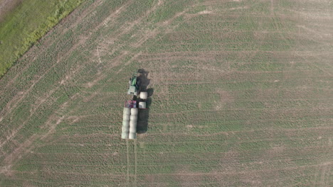 aerial top view of green industrial tractor machine loading and hauling hay bales in rural flat plains farmland and countryside, saskatchewan, canada, rising directly above drone