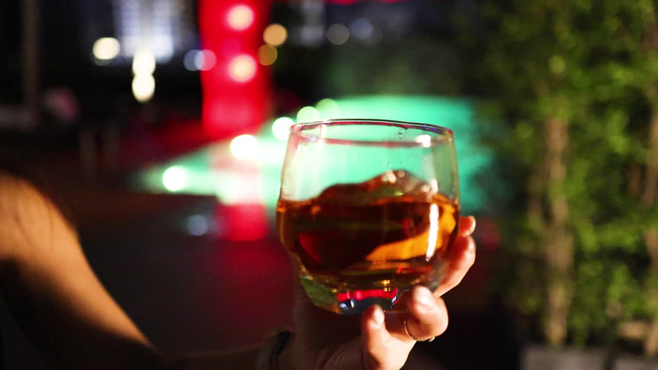 A person swirls a cocktail in a clear glass, illuminated by colorful nightlife lighting, with a blurred outdoor bar or lounge setting in the background