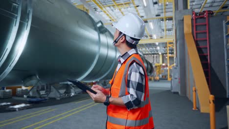 vista lateral de un ingeniero masculino asiático con casco de seguridad mirando la tableta en su mano y mirando a su alrededor mientras está de pie en una fábrica de fabricación de tubos