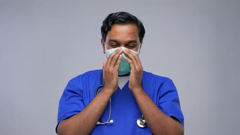 Indian-Male-Doctor-in-Blue-Uniform-Putting-Mask-on.healthcare,-profession-and-medicine-concept-happy-smiling-indian-doctor-or-male-nurse-in-blue-uniform-putting-on-face-protective-medical-mask-for-protection-from-virus-disease-over-grey-background