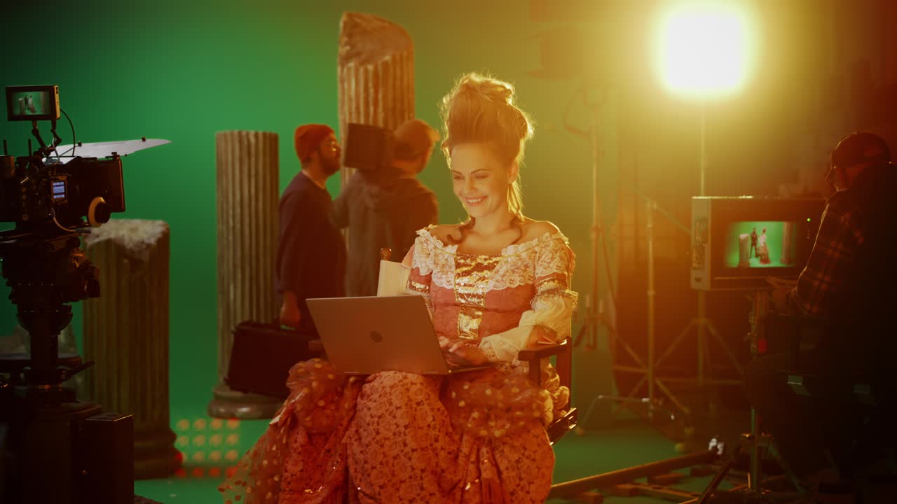 en el set de la película de drama de vestuario de época: hermosa actriz sonriente con un vestido renacentista, sentada en una silla usando una computadora portátil con pantalla verde en el fondo. estudio zoom en