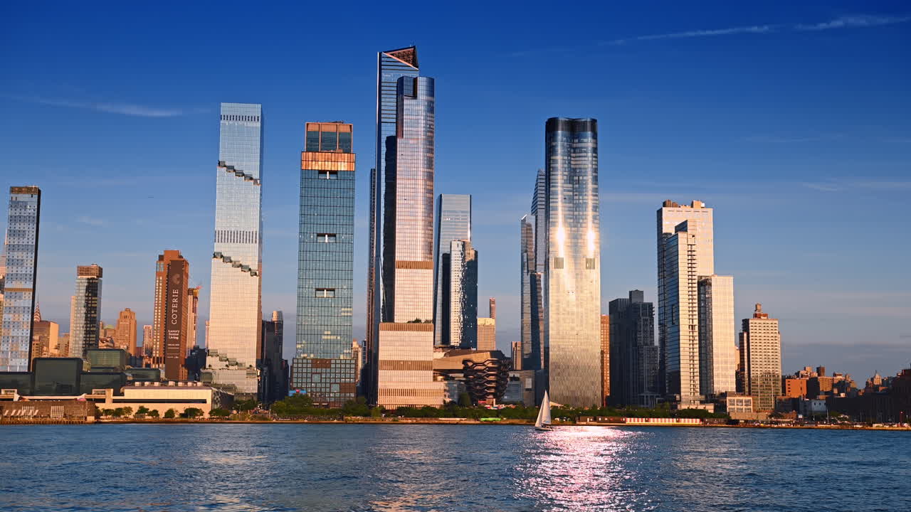 New York, USA, 1 August 2025: Glass skyscrapers reflecting sunlight. Vessel Structure at the waterfront. A sailboat moves close to the shore. View from the riverscape