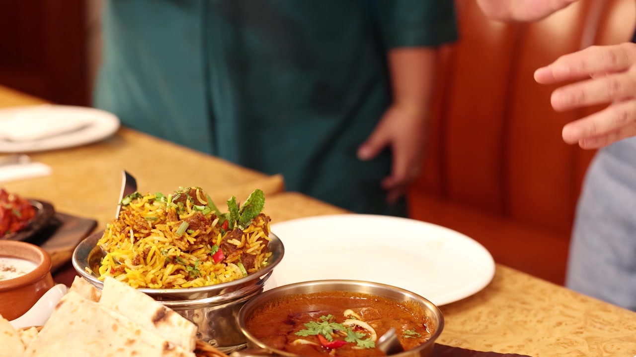 Statisk mellembillede af forskellige indiske retter på et restaurantbord med folk, der gestikulerer i nærheden.