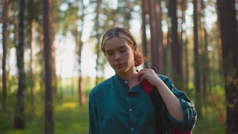 young woman walking through peaceful forest, carrying red backpack over her shoulder, adjusting strap while gazing thoughtfully into distance, sunlight casts warm glow on her face