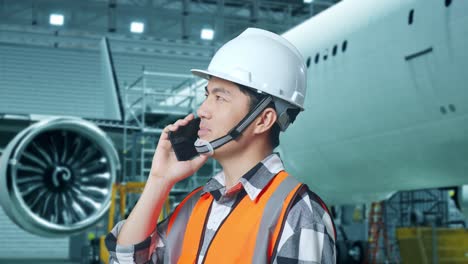 vista lateral cercana de un ingeniero masculino asiático con casco de seguridad hablando en teléfono inteligente mientras está de pie con el avión en el hangar