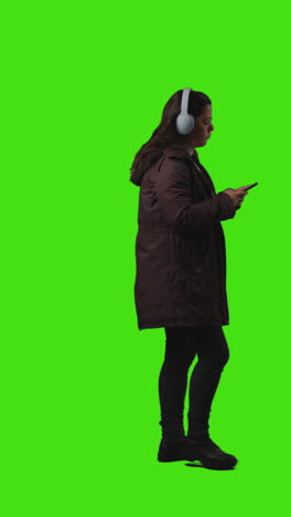 vertical video full length shot of woman wearing wireless headphones streaming music from mobile phone walking across frame against green screen 1