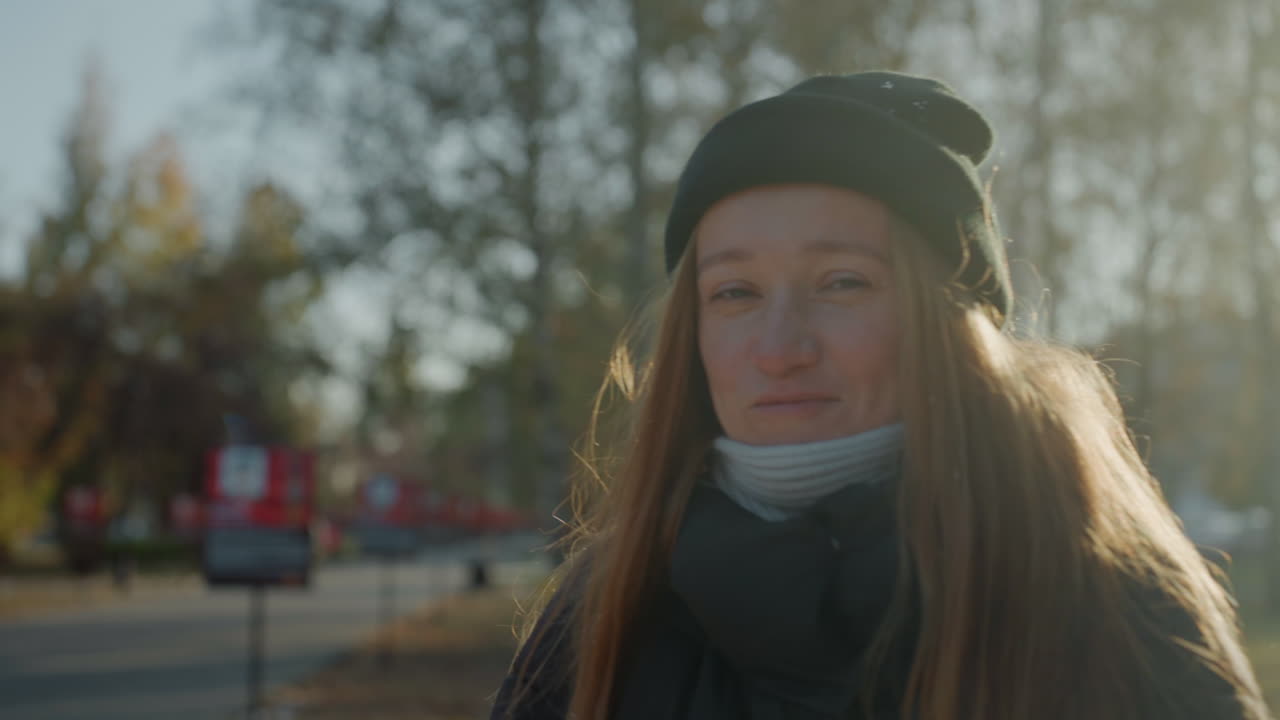 Smilende kvinde i solbeskinnet park. Oprigtigt efterårsportræt med gyldent modlys, strikhue og tørklæde, langt hår der fanger lyset, varme fregner og afslappet blik, venlig atmosfære og naturlig livsstil.