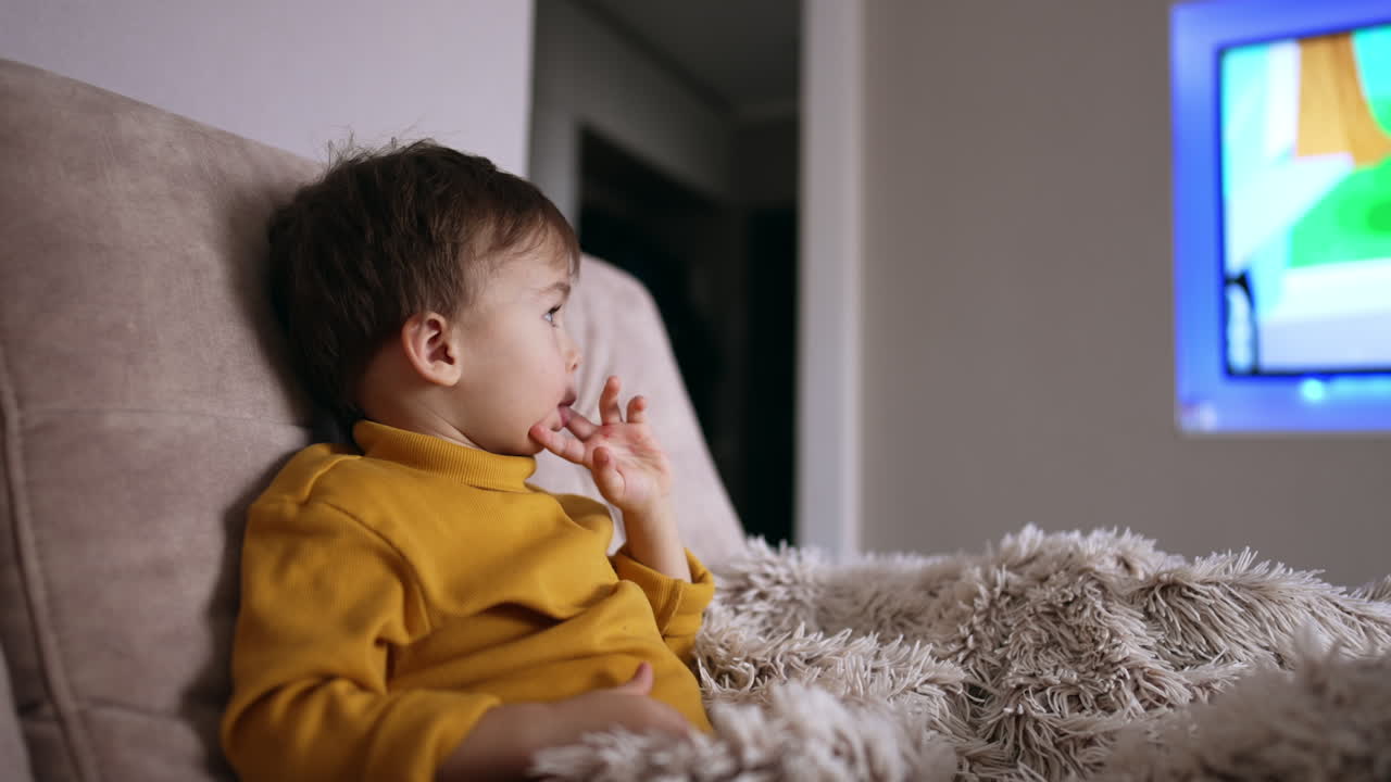 Relaxed baby boy sits on the sofa holding a finger in his mouth. Cute smiling toddler watching TV.