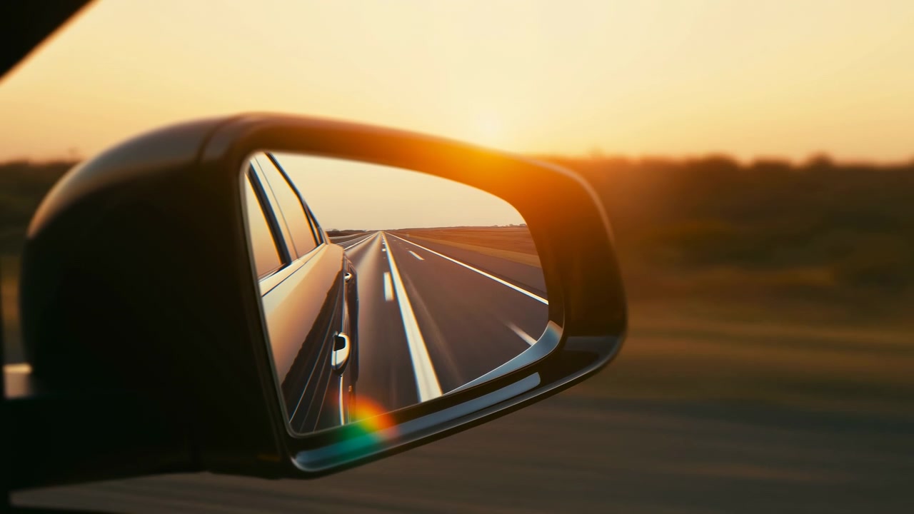 Vista ravvicinata dello specchietto retrovisore lato passeggero che riflette un'autostrada dritta a due corsie, linee di corsia convergenti, un cielo caldo al tramonto, un orizzonte lontano, un leggero flare dell'obiettivo, il lato dell'auto, un campo a bordo strada e una sera mite.