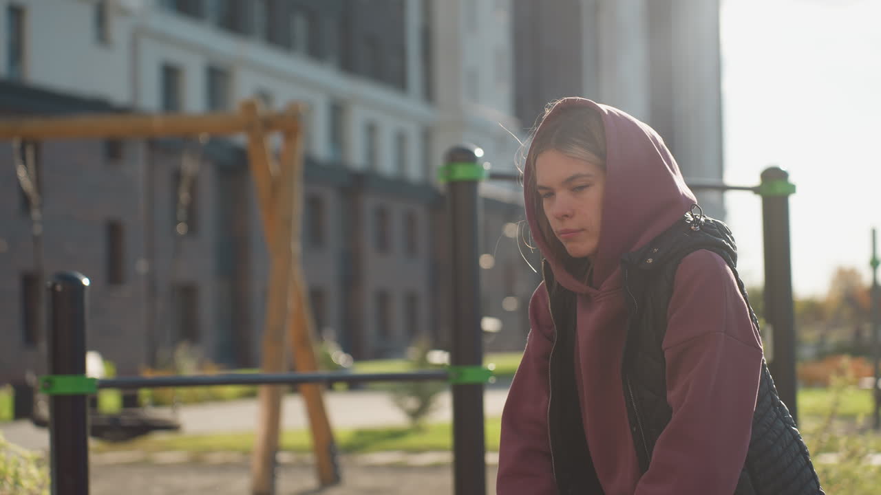 Outdoor fitness trainer in hoodie and quilted vest lunges forward resting hands on bent knee under sunset glare against urban park buildings backdrop showing relief on face