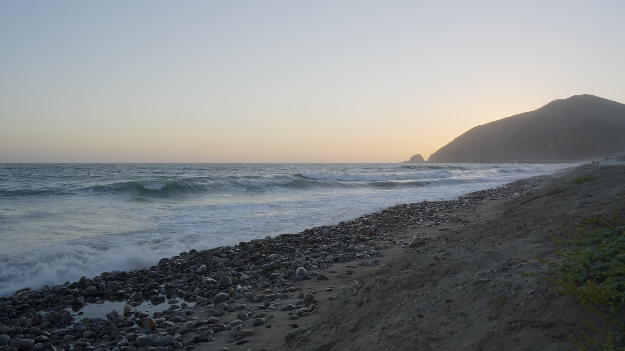 toma estacionaria de olas rompiendo en las orillas de la playa de mondo con la puesta de sol detrás de una montaña en el fondo ubicada en el sur de california