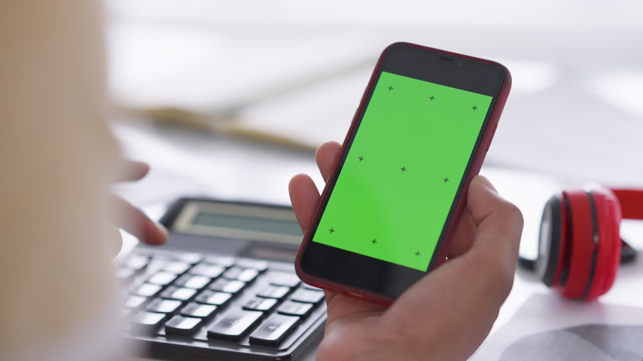 Close-up chromakey smartphone in male African American hand with fingers pressing keys on calculator at background. Unrecognizable young man accounting business profitability in home office.
