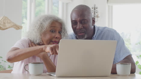 senior couple sitting around table at home shopping or booking holiday on laptop