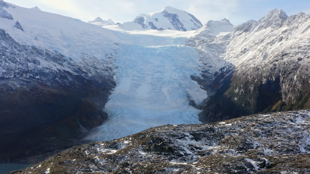 Glacier ice descends into fjord water valley with mountain rocky ridges as sunlight illuminates hills and casts dark shadows