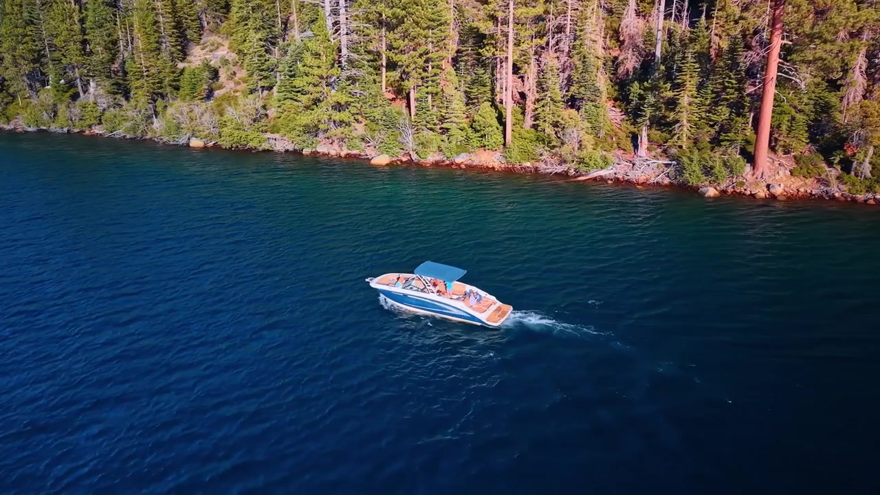 Motorboat cruising beside the forested shore of Emerald Bay. A varnished boat carries passengers across blue water, leaving white foam below a line of conifers.