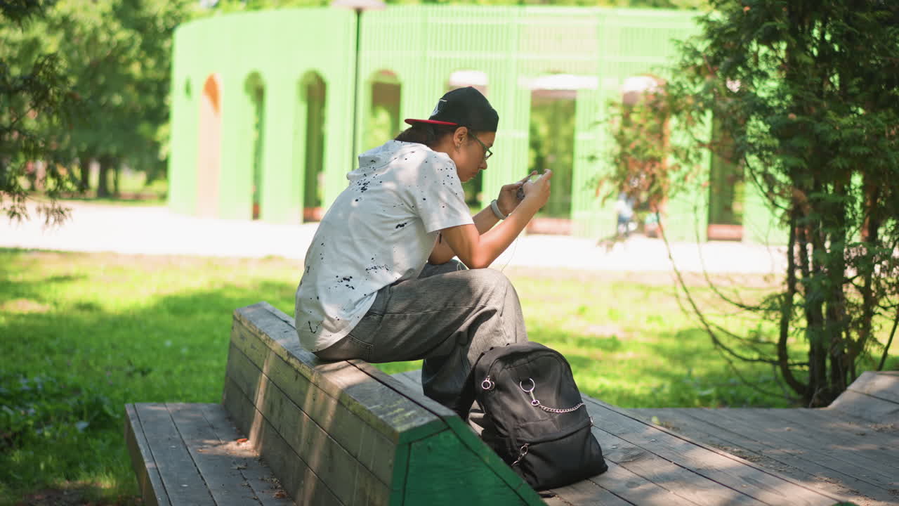 Joven sentado en un banco, estudiante universitario absorto en el móvil, mochila a sus pies, luz solar moteada entre los árboles, fondo bokeh suave, gorra informal y camisa estampada, ambiente veraniego tranquilo, concentrado