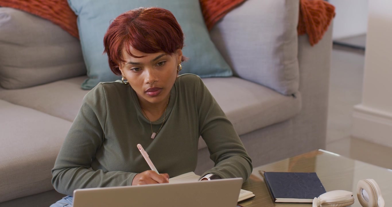 African American woman transcribing from laptop, writing notes on notebook with pink pen at home