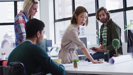 Diverse-office-colleagues-wearing-face-masks-discussing-over-blueprint-plan-at-modern-office