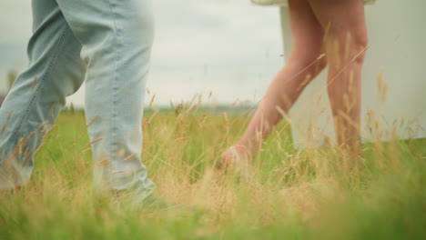 a side shot of two people walking through a grassy field. the man wears jeans and black shoes, while the woman, in a short gown, walks beside him. the focus is on the legs moving through the nature