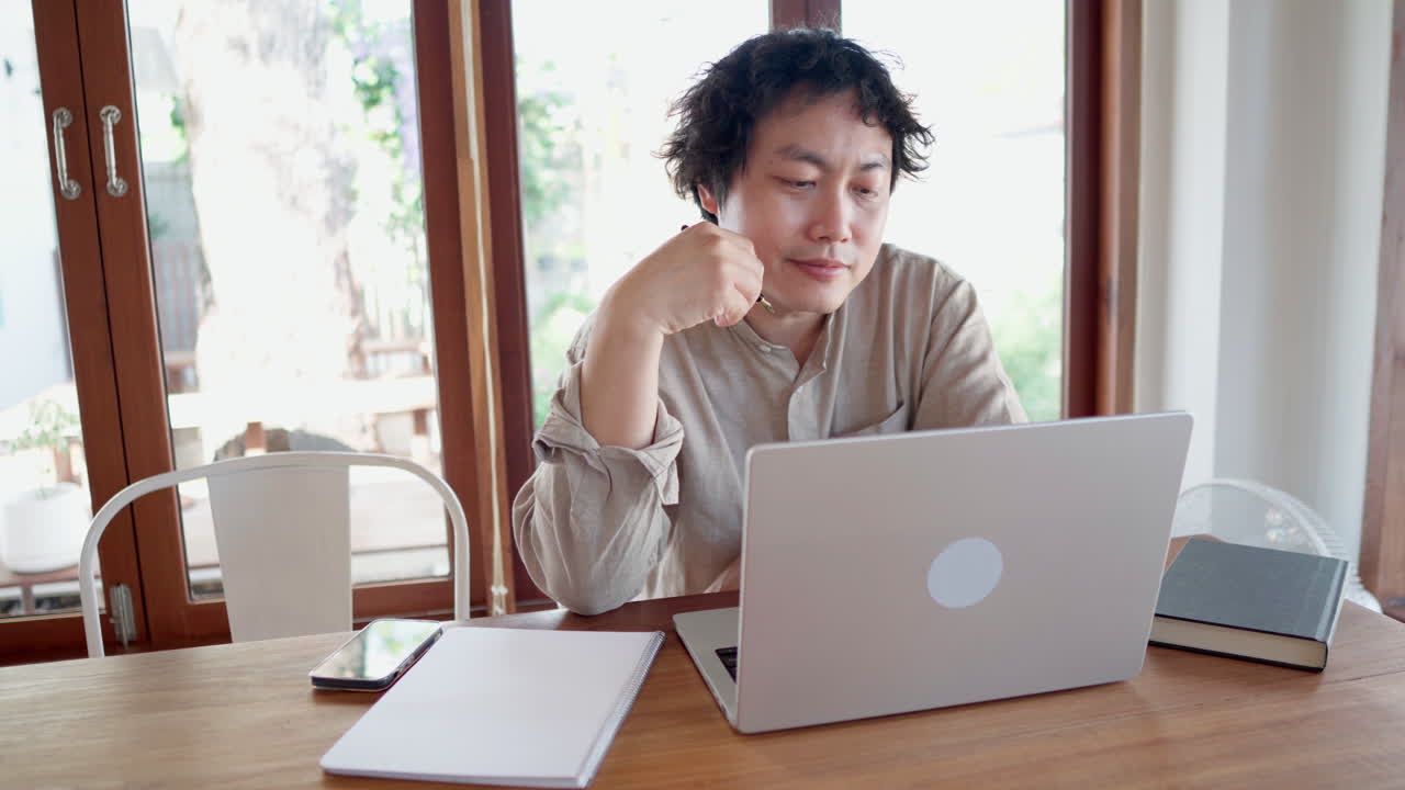 집중된 심각한 아시아인 노트북에서 온라인 학습 과정을 보고 메모를 취합니다.