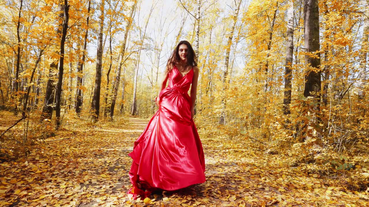 A stunning woman in a flowing red gown walks confidently through an enchanting golden forest, surrounded by autumn leaves that create a magical atmosphere of beauty and grace