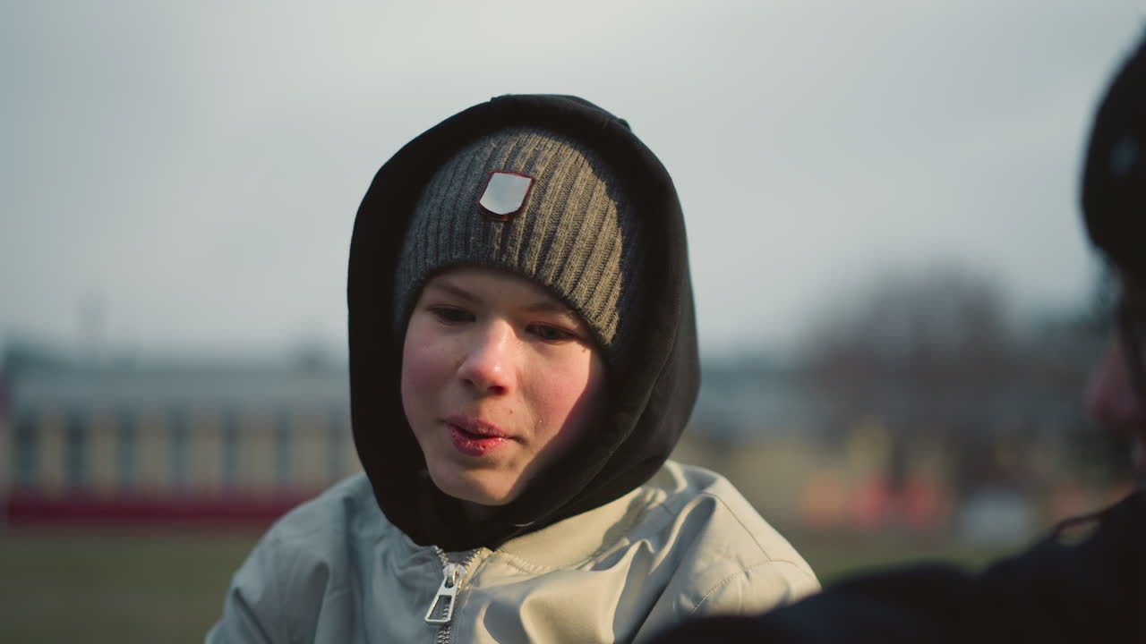 Close-up of a young boy wearing a gray top, beanie and a black hoodie, engaged in a discussion with someone, with a blur background of building