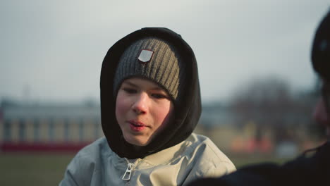 close-up of a young boy wearing a gray top, beanie and a black hoodie, engaged in a discussion with someone, with a blur background of building