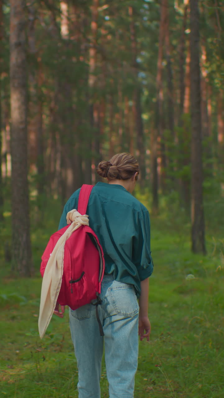 Back view of woman walking through lush forest, holding red backpack, dressed in green shirt, blue jeans, and white canvas shoes, touching tall grass strands along the forest trail