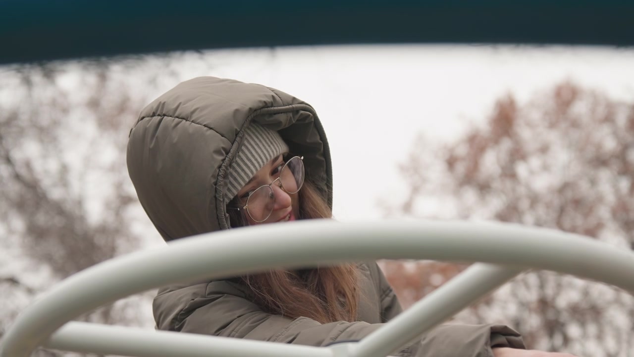 Woman with glasses smiling in winter park. Close shot of joyful woman wearing hood and beanie outdoors. Smiling woman with glasses and knit hat enjoying peaceful winter scene in park