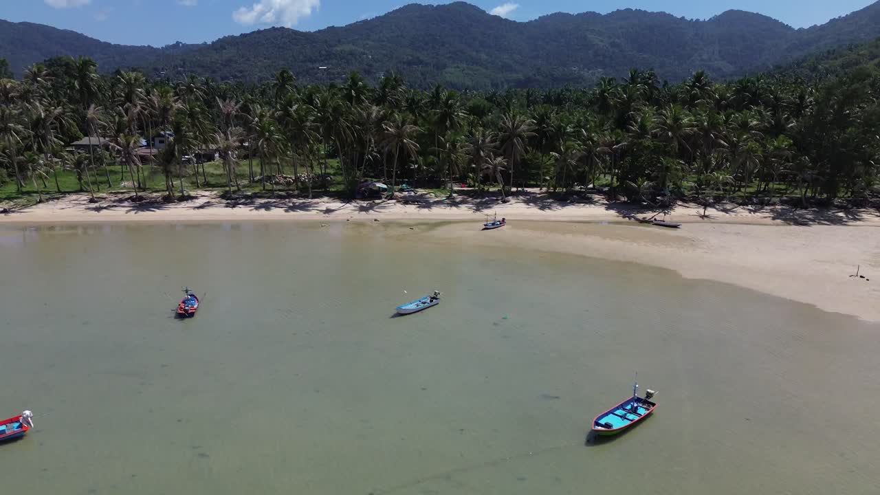 Stunning drone footage captures traditional Thai fishing boats resting on the clear, shallow waters off Koh Phangan island in Thailand. A serene tropical landscape island scenery