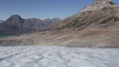 Low-altitude-aerial-view-from-drone-flying-over-ice-field-of-glacier-in-Greenland-with-rugged-terrain-of-steep-mountain-slopes-and-jagged-peaks-reaching-up-to-clear-blue-sky