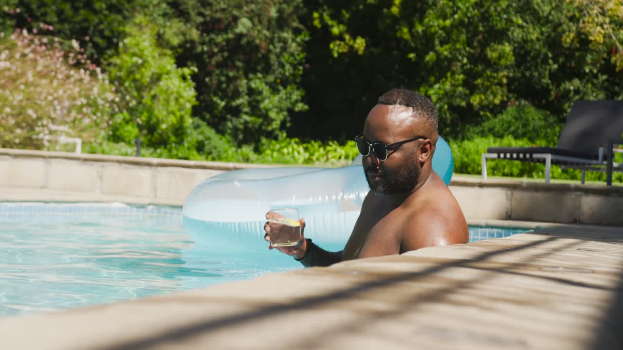 African American shirtless man noticing camera, turning and toasting with glass in pool, copy space