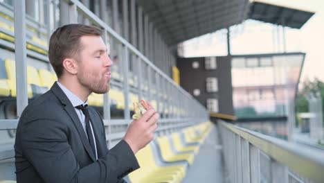 Young Attractive Businessman Manager Sitting in Sports Arena and Eating Hot Dog. Young attractive businessman, manager sitting in sports arena and eating hot dog. Fast food. Harmful food. Office worker’s lunch