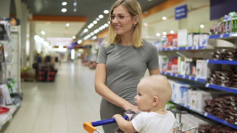 Young-mother-with-her-little-baby-sitting-in-a-grocery-cart-in-a-supermarket-is-pushing-the-cart-forward-walking-among-different