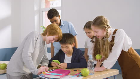 Estudiantes-Multiétnicos-Se-Agrupan-Alrededor-De-Un-Escritorio-Donde-Un-Estudiante-Está-Sentado-Usando-Un-Teléfono-Inteligente-En-El-Aula-De-Inglés