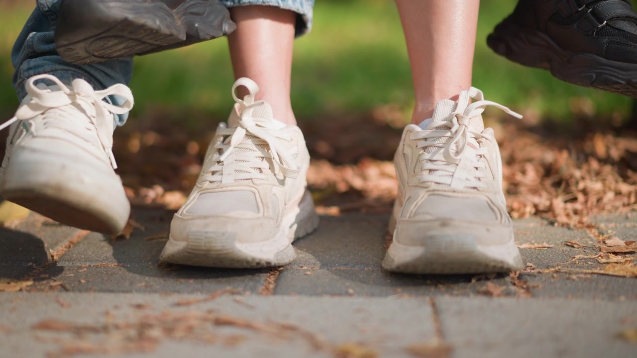 Nærbillede af to par fødder i bevægelse, det ene par iført hvide sneakers og det andet sorte sko, på en brolagt sti med faldne blade. Scenen fanger subtil fodbevægelse mod en udendørs baggrund.
