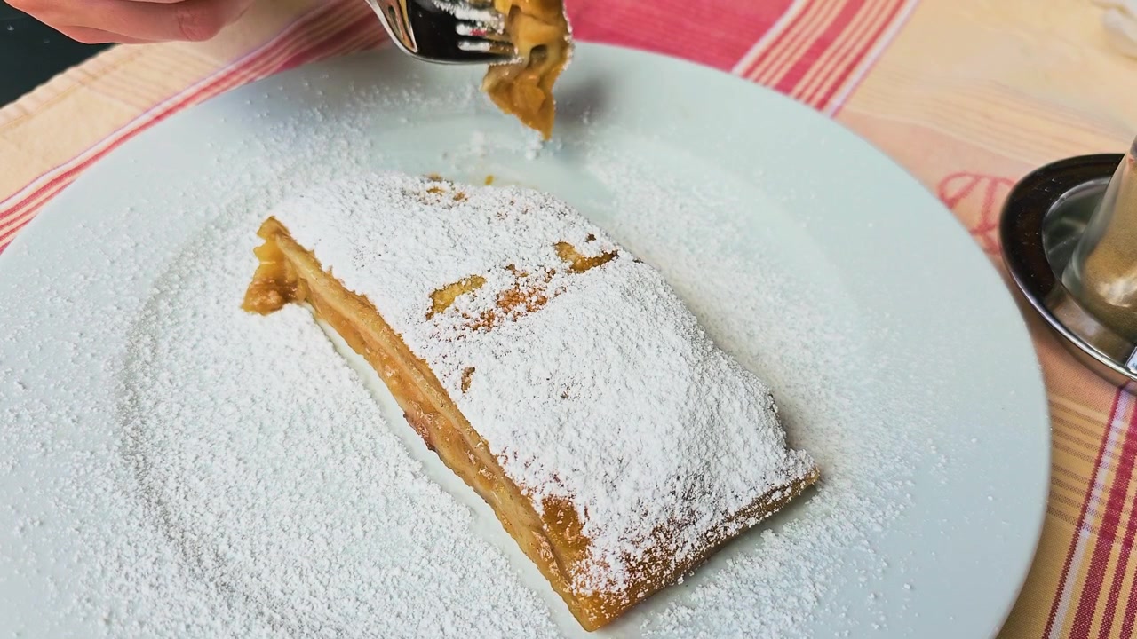 Beyaz bir tabakta pudra şekeri serpilmiş Avusturya elmalı strudel'e batan çatal, açık hava Avrupa kafe restoran masasında tatlı elma dolgulu pul pul dökülen hamur işi, tatlı yakın çekim.