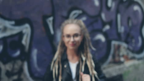 Close-Up-Of-The-Camera-Zooming-In-The-Face-Of-The-Pretty-Stylish-Girl-In-Glasses-And-With-Dreadlocks-Standing-On-The-Graffity-Wall-Background-And-Smiling