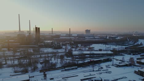 Vista-Aérea-De-La-Zona-Industrial-Cubierta-De-Nieve,-Con-Varios-Edificios-Y-Estructuras-Bajo-Un-Cielo-Dorado-Al-Amanecer.