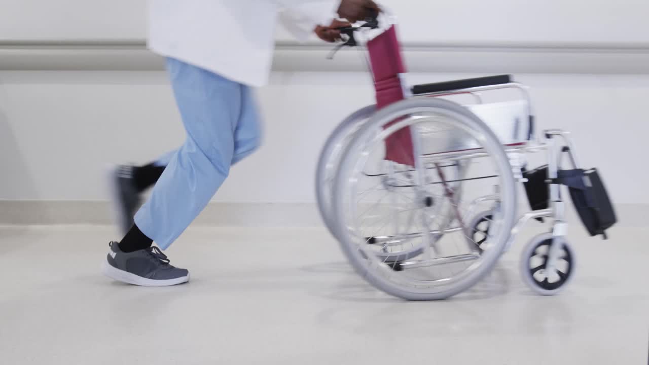 Lowsection of african american male doctor running with wheelchair in hospital corridor, slow motion