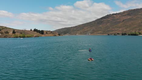 Cámara-Lenta---Aérea---Personas-Con-Moto-Acuática-Y-Galletas-En-El-Lago-Dunstan-Cerca-De-La-Presa-Clyde,-Otago-Central,-Nueva-Zelanda-Con-Montañas-Y-Nubes-En-El-Fondo