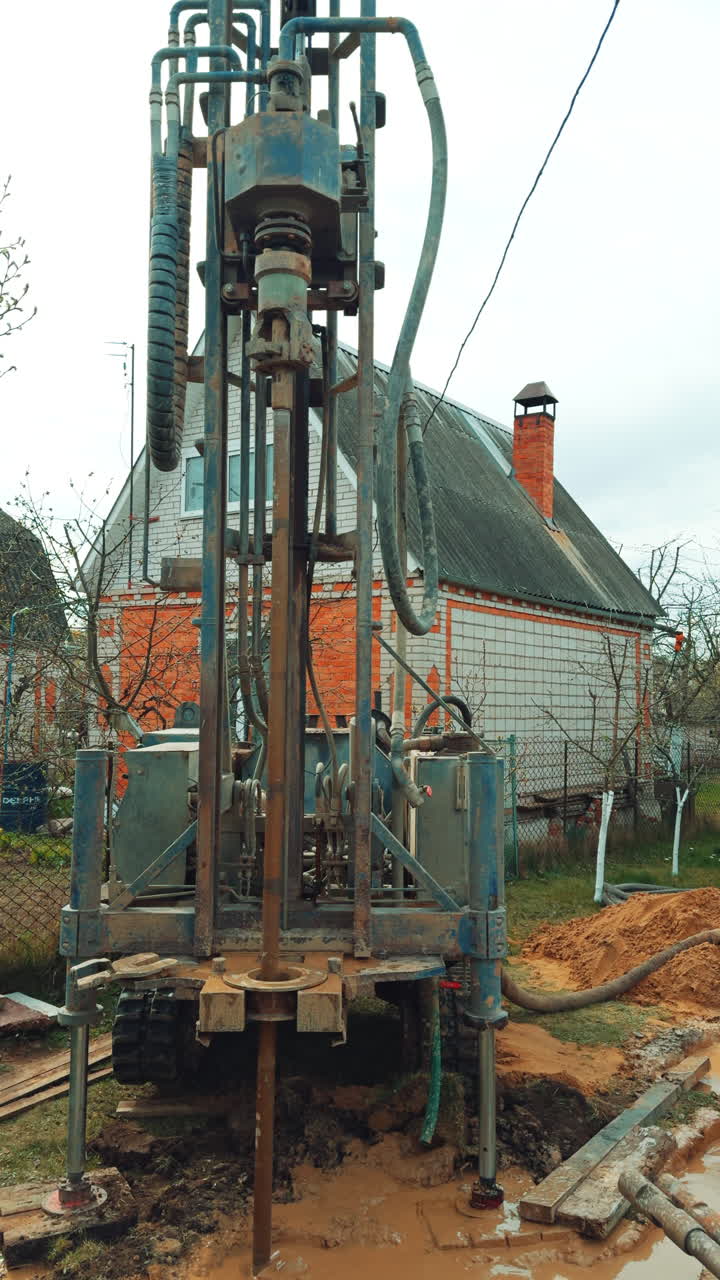Boring the well with the help of special machinery. Drilling equipment producing the water bore. Private cottage at backdrop. Vertical video.