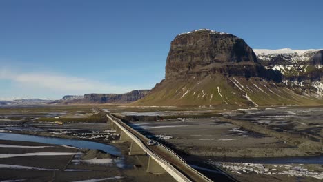 Luftaufnahme-Des-Autos,-Das-Die-Lomagnupur-Brücke-In-Island-überquert,-Mit-Massivem-Berg-Im-Hintergrund---Schöner-Sonniger-Tag-Mit-Blauem-Himmel-Auf-Der-Insel