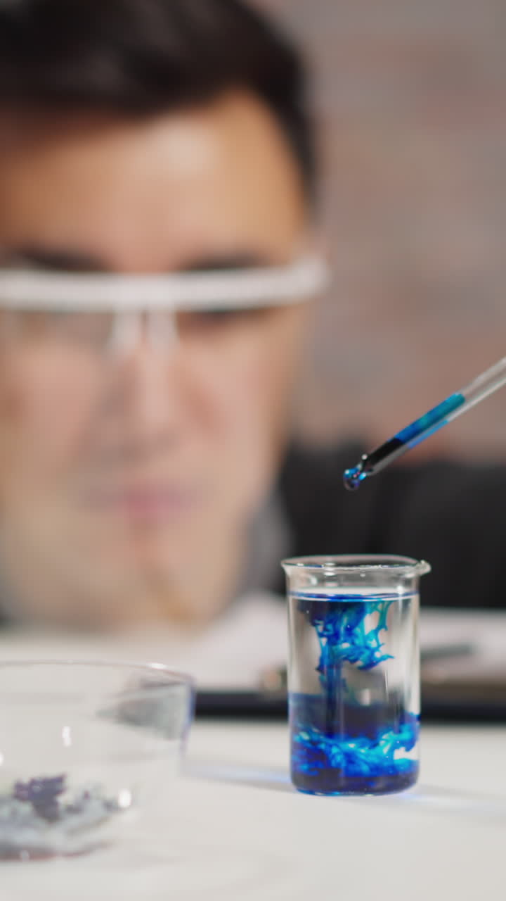 Skilled scientist in protective gloves and shield adds dark blue liquid into flask with clear water doing experiment in medical lab closeup