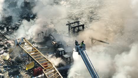 Eine-Umlaufende-Luftaufnahme-Zeigt-Eine-Gruppe-Von-Feuerwehrleuten-Auf-Einer-Leiter,-Die-An-Einem-Sonnigen-Tag-Brennende-Fahrzeuge-Nach-Einer-Gasexplosion-Löschen