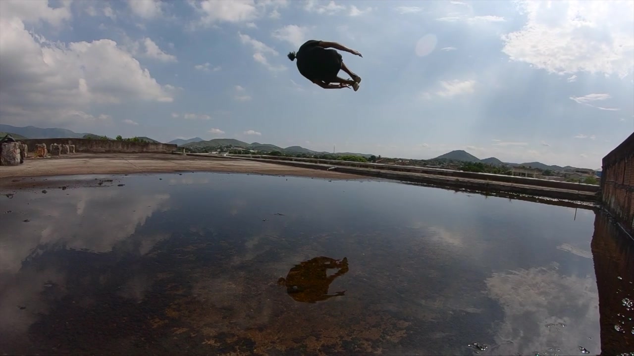 Video al rallentatore di un giovane freerunner che esegue un sideflip, creando un effetto specchio in una pozzanghera sul tetto di un edificio abbandonato.
