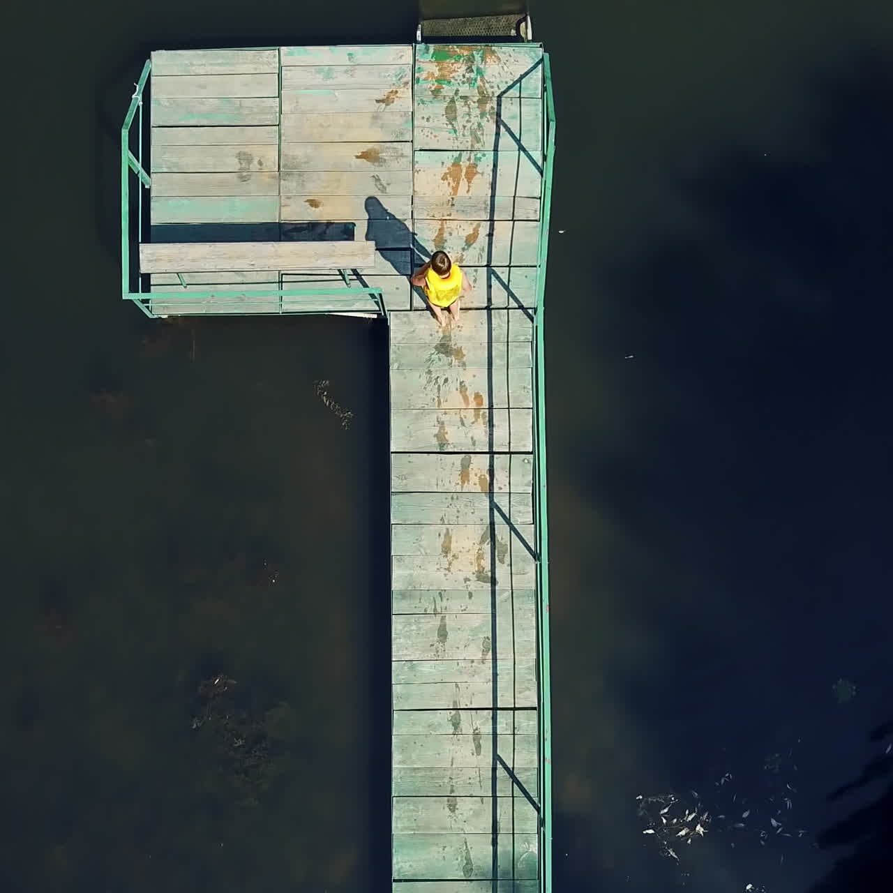 Little boy walking on the long wooden bridge and jumping into the water in a sunny day. Kid wearing waistcoat having a good time in the river on his summer holidays. Aerial view