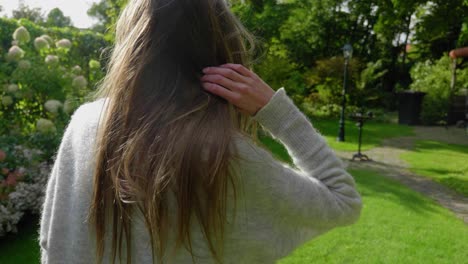 Slow-motion-shot-following-a-young-attractive-woman-walking-through-a-lush-green-garden,-arranging-her-hair-while-strolling-casually