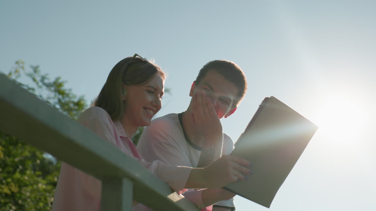 분홍색 을 입은 여자가 미소 짓고 그녀의 형에게 그녀의 태블릿에서 무언가를 보여주면서 머리를 흔들고, 한 손으로 태블릿을 가리키고 다른 손으로 입을 고 있습니다.