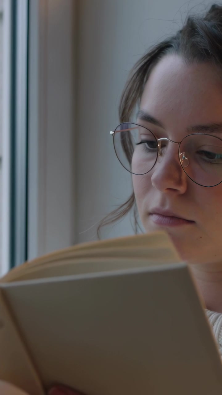 Video vertical: Mujer con gafas y suéter sosteniendo un libro de bolsillo, mirando por la ventana y pasando una página.